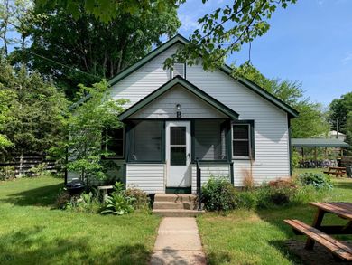 Small white house with green trim and a front porch, surrounded by a sunny yard and trees