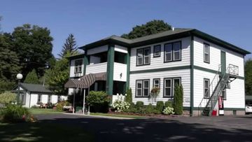 White two-story house with green trim, front porch, and driveway in a sunny residential setting