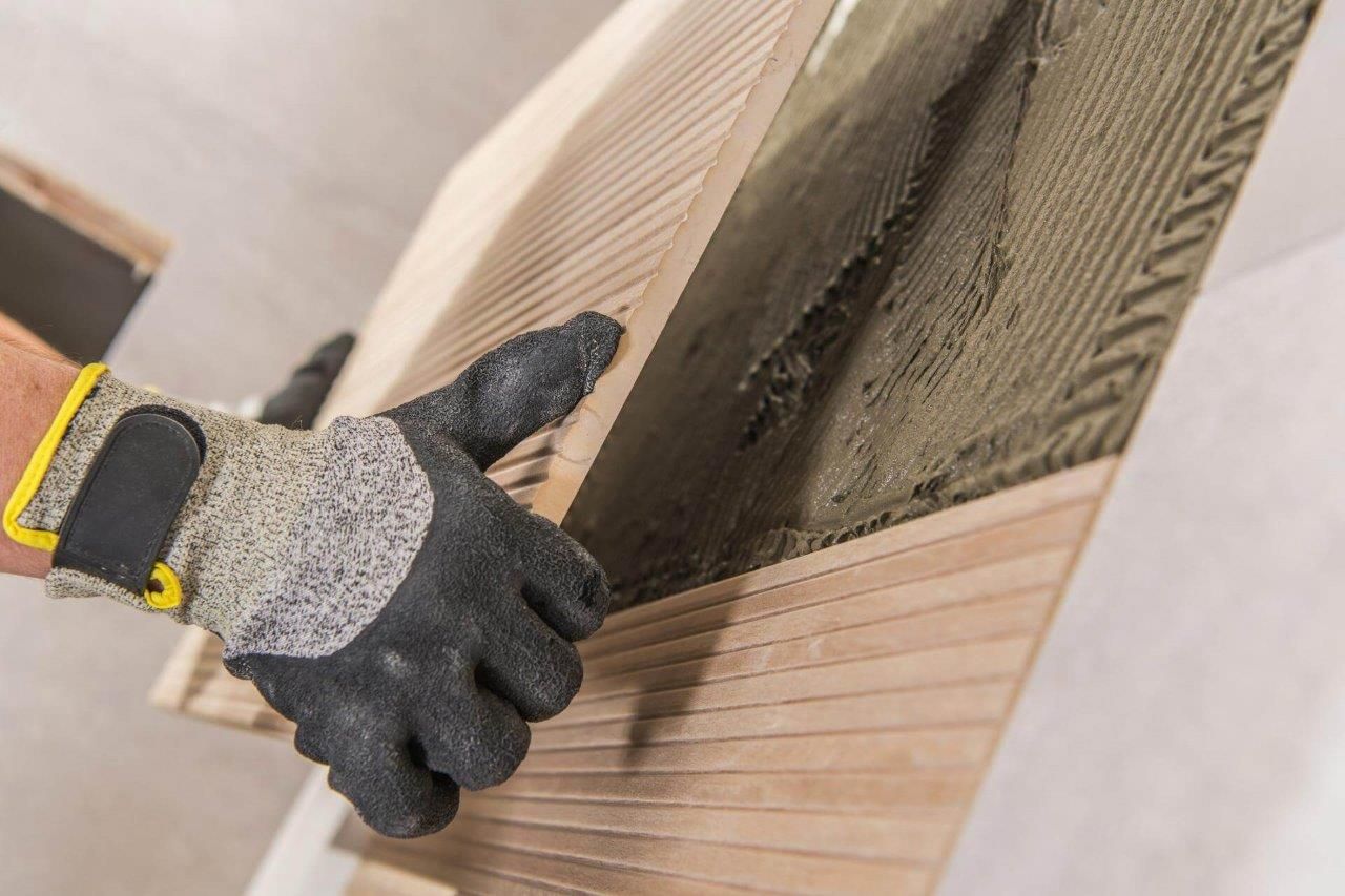 Persona che indossa guanti da lavoro mentre installa una piastrella di legno con adesivo su un muro.