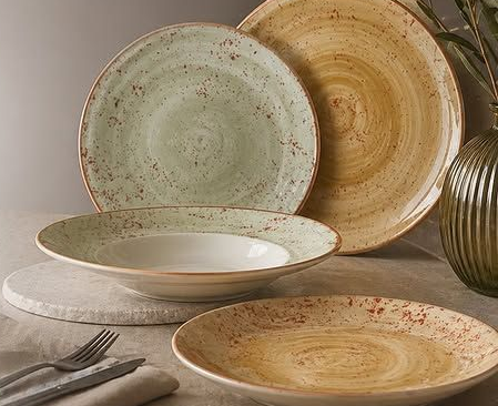 Piatti e ciotole in ceramica impilati, nei toni tenui del verde, del beige e del marrone, su una tavola apparecchiata.