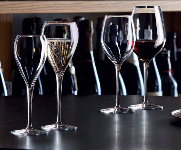 Calici da vino e un flûte da champagne su un bancone scuro, con bottiglie sfocate sullo sfondo.
