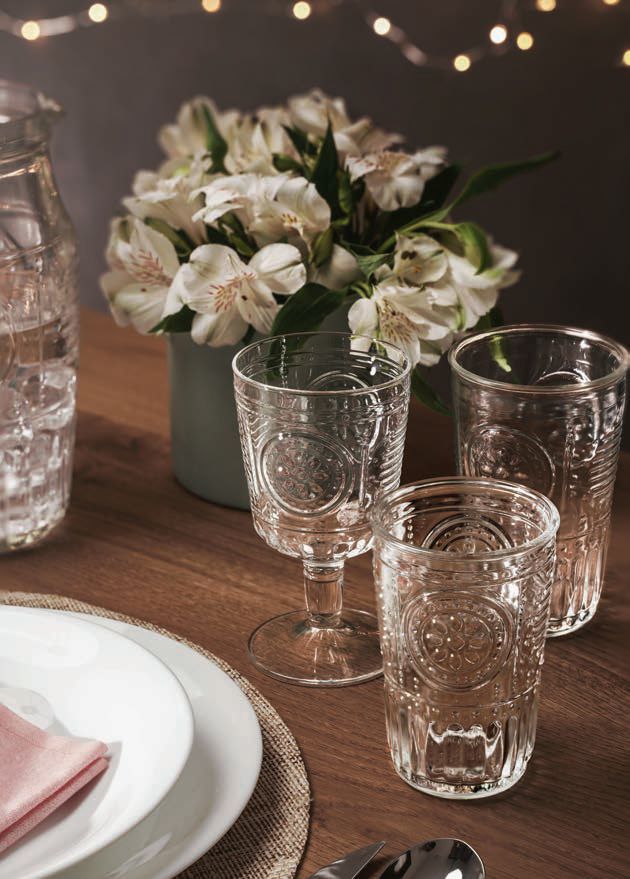 Tavola apparecchiata con fiori bianchi in vaso, bicchieri trasparenti e un tavolo di legno illuminato da candele.