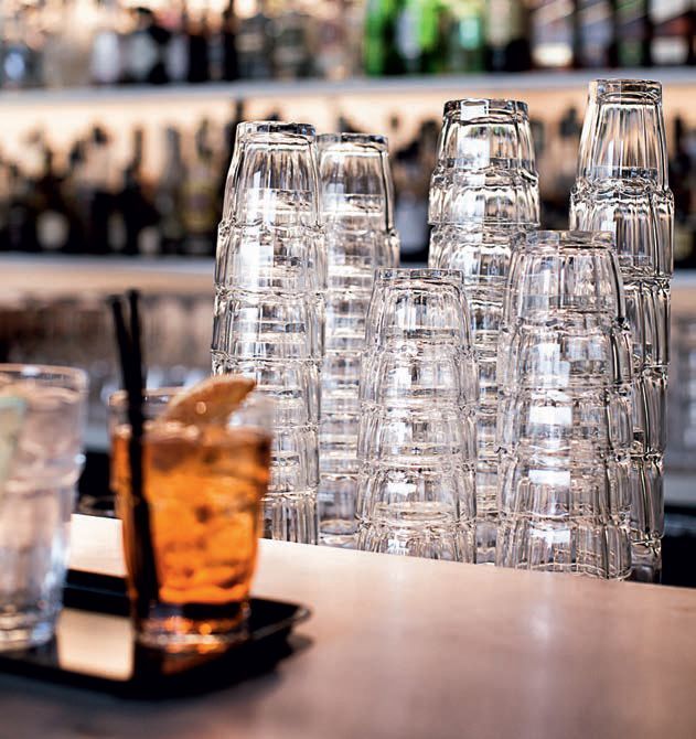 Bicchieri trasparenti impilati sul bancone di un bar, accanto a un cocktail ambrato con cannuccia.