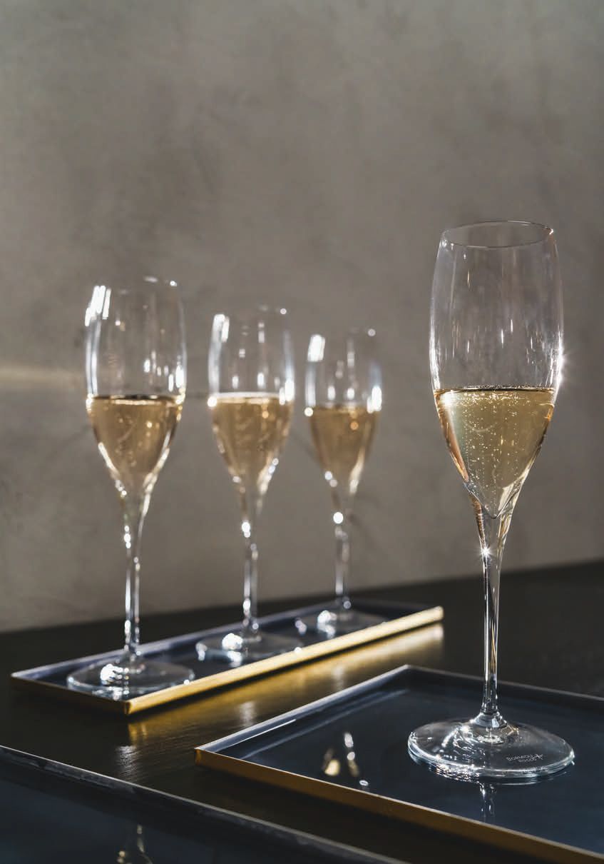 Quattro flûte da champagne su vassoi a specchio contro una parete grigia, con un bicchiere più grande davanti.