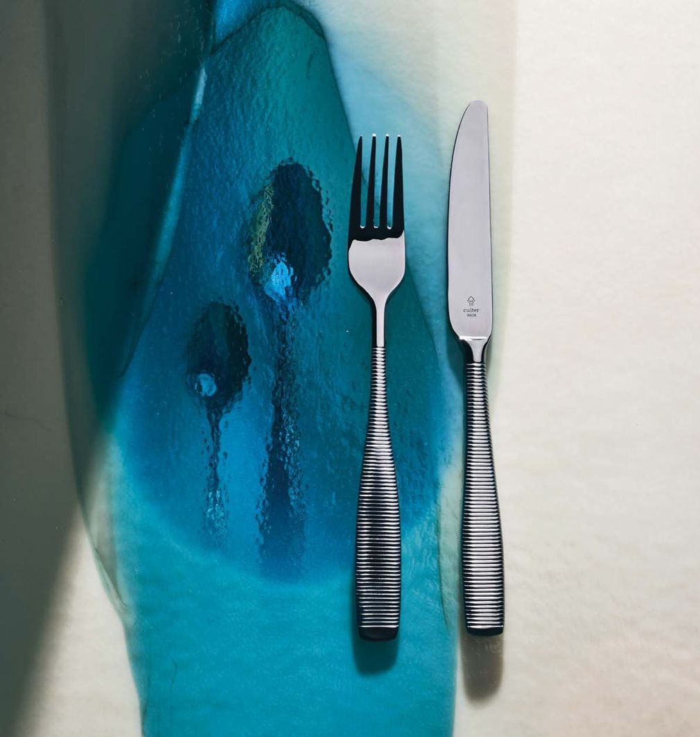 Piatto di vetro blu con forchetta e coltello su un tavolo luminoso, con un riflesso simile a un volto sul piatto.