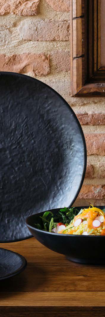 Una ciotola nera di insalata su un tavolo di legno accanto a un grande piatto scuro, appoggiato a un muro di mattoni.