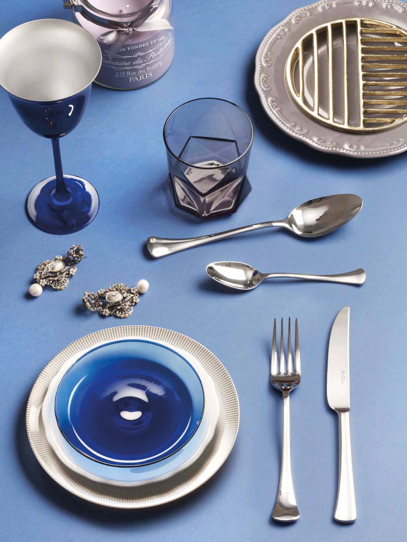 Apparecchiatura da tavola blu e argento con piatti, bicchieri, posate e anelli portatovaglioli decorativi su una tovaglia blu.