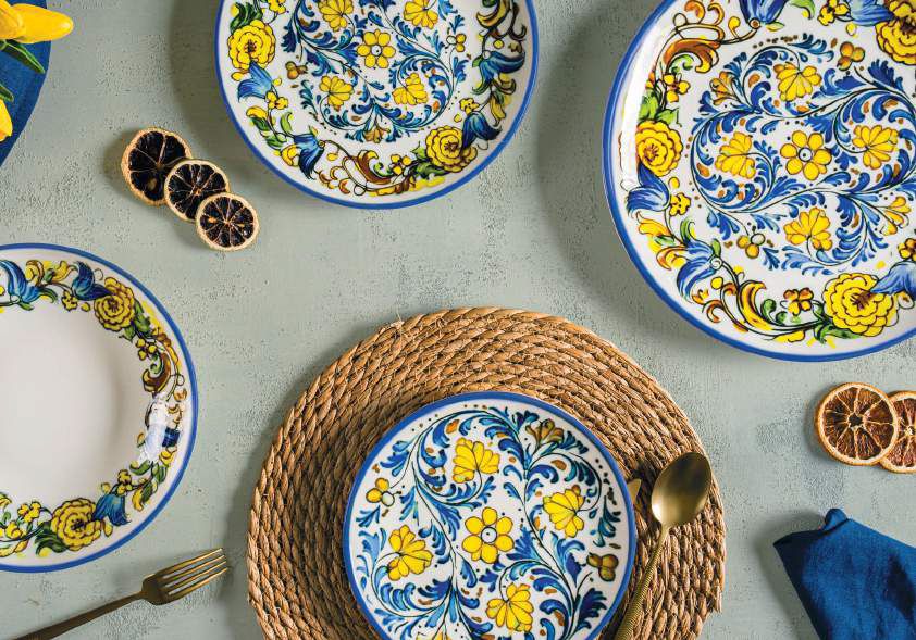 Piatti floreali blu e gialli su un tavolo con tovaglietta intrecciata, limoni e tovagliolo, in una mise en place elegante.