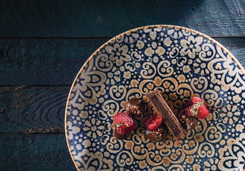 Piatto blu decorato con un piccolo dessert e frutti di bosco rossi su un tavolo di legno scuro.