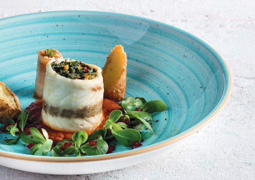 Piatto gourmet impiattato con filetto di pesce arrotolato, verdure e guarnizione croccante in una ciotola turchese.