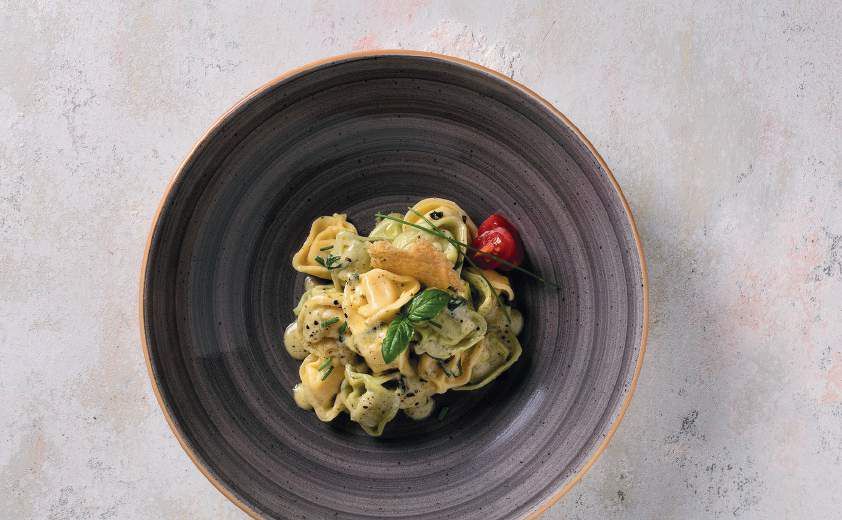 Pasta con salsa cremosa e basilico su un piatto scuro, guarnita con un pomodorino e erbe aromatiche.