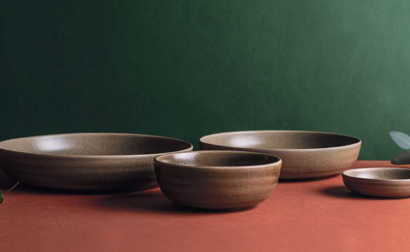 Quattro ciotole di ceramica nei colori marrone, beige e rosso, disposte su un tavolo su uno sfondo verde scuro.