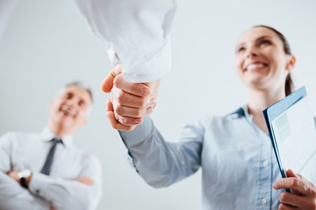 Woman shaking hands with a person, smiling, holding a folder. Man watches in background.