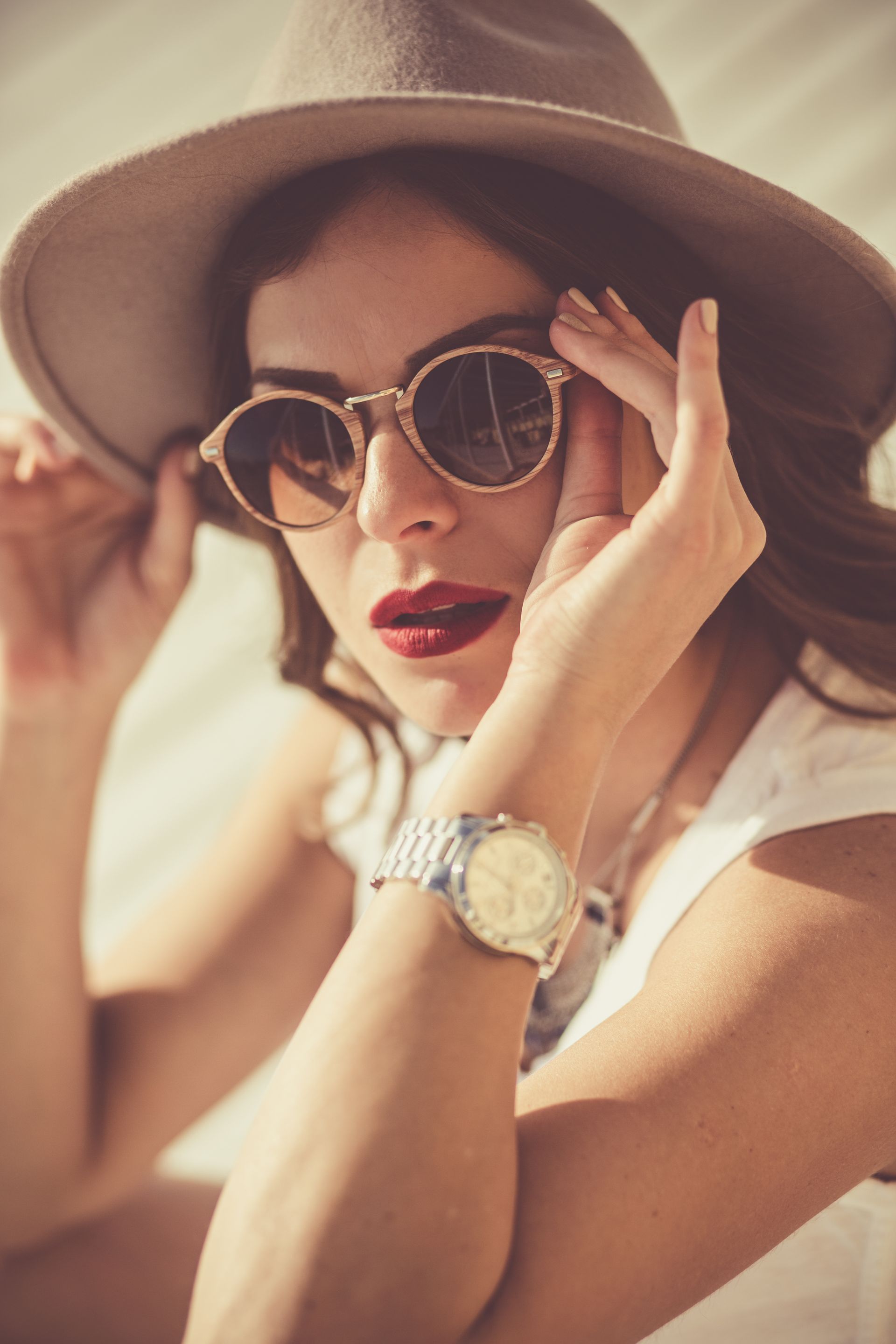 Woman in hat and sunglasses adjusting eyewear, wearing a watch, with bright red lipstick.