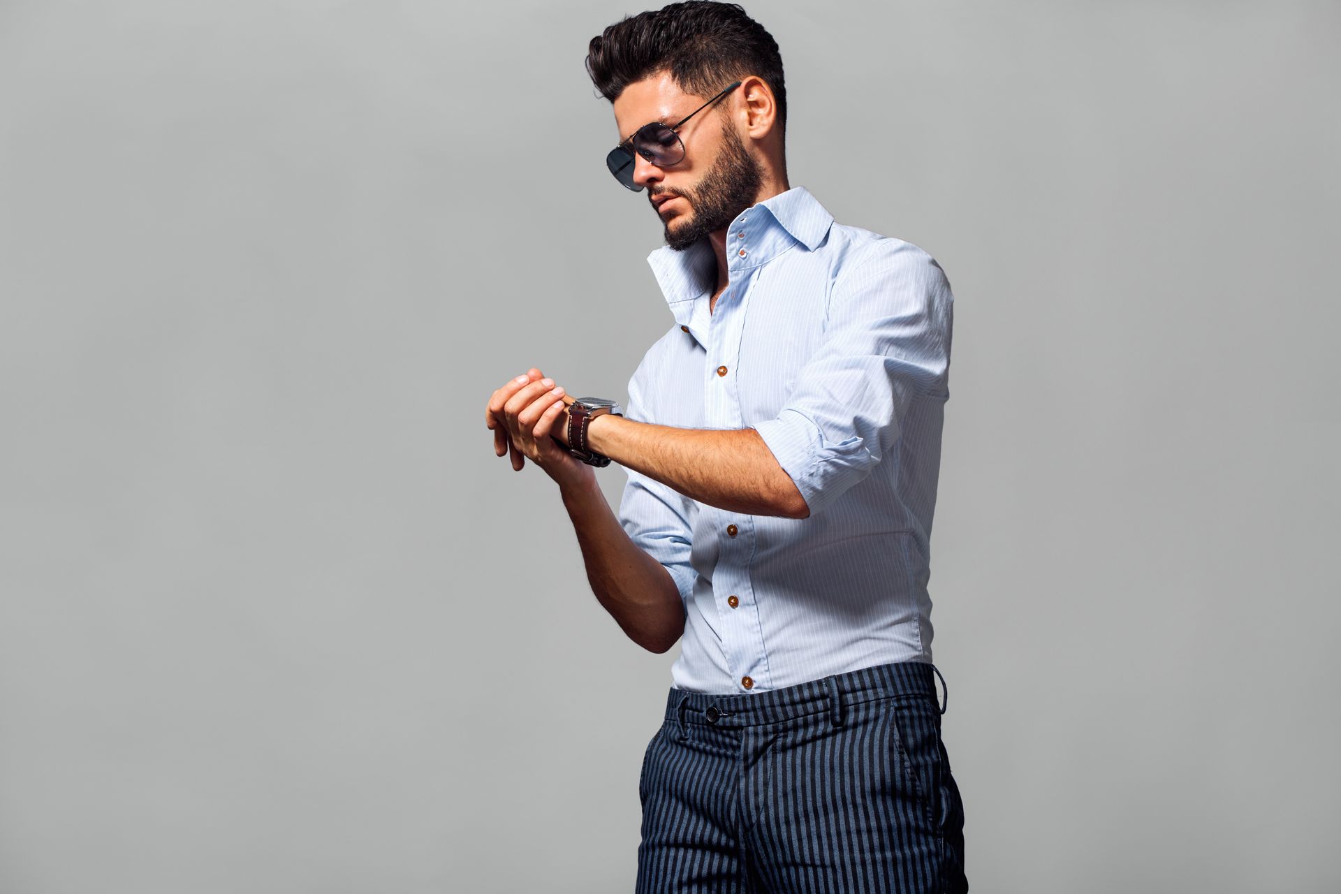 Man in sunglasses checks his wristwatch, wearing a light blue dress shirt and striped pants.