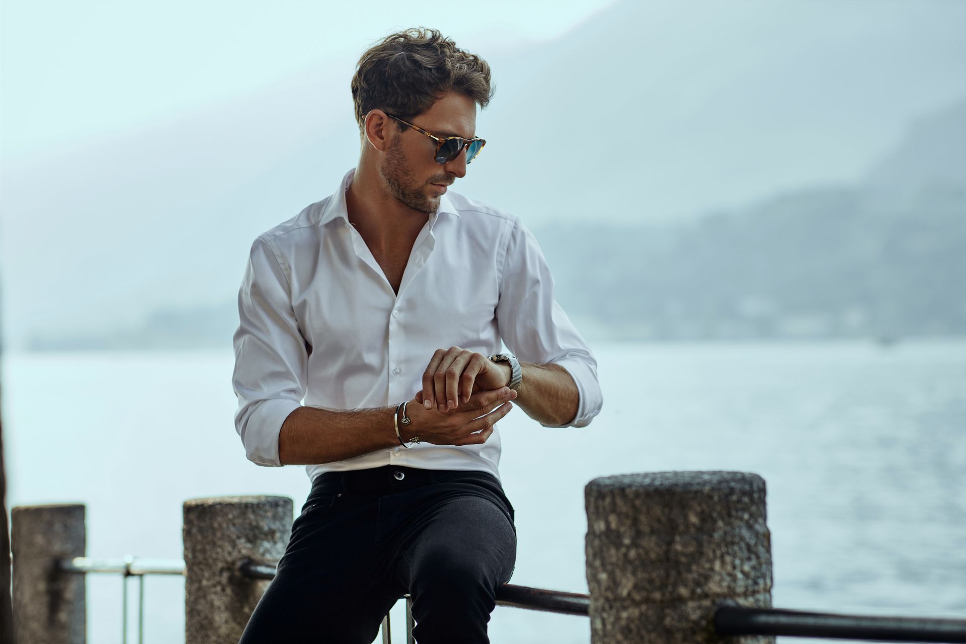Man in white shirt and sunglasses looking at his watch by a lake.