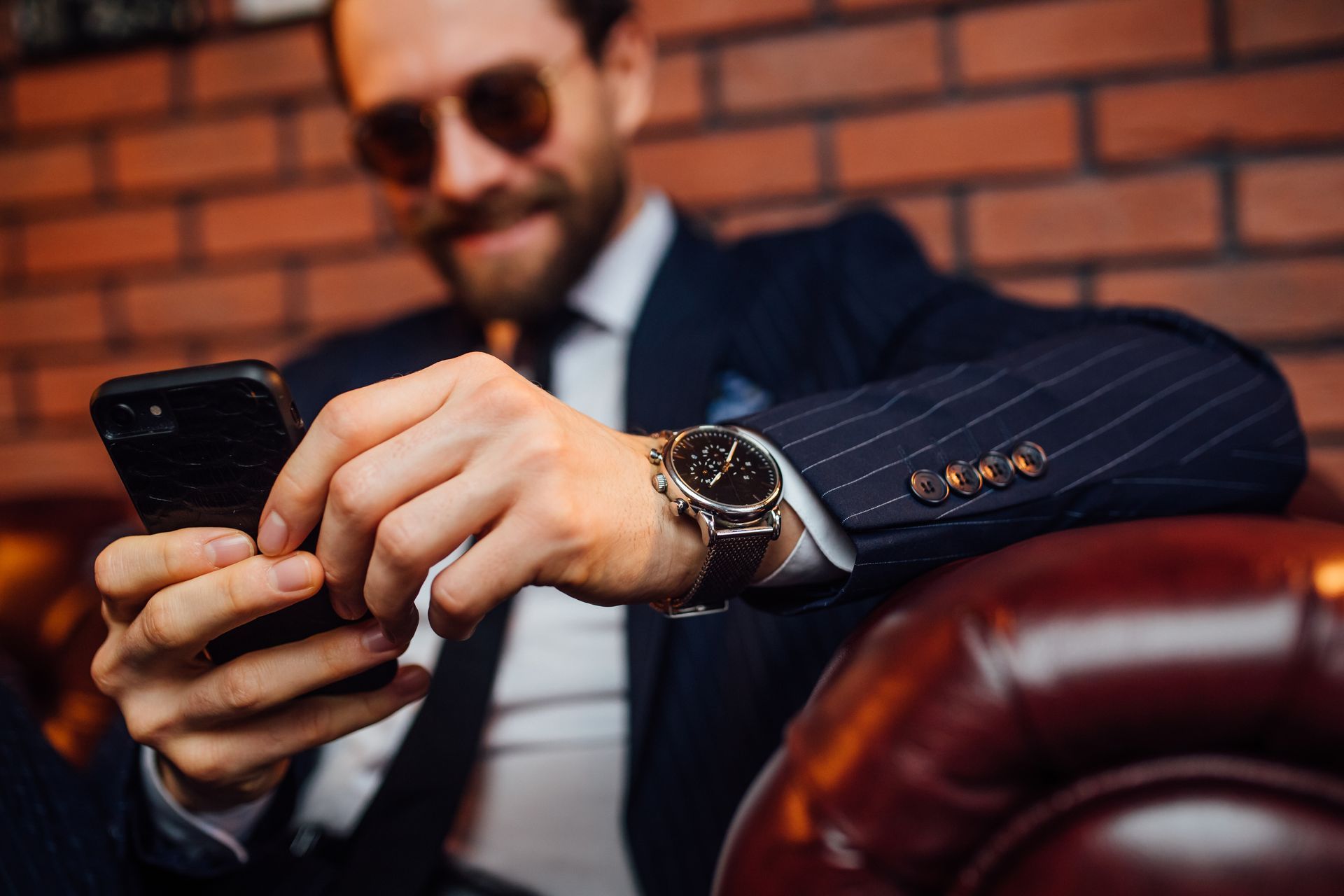 Man in suit and sunglasses using a phone, wearing a watch. Leaning on a leather chair, brick wall backdrop.