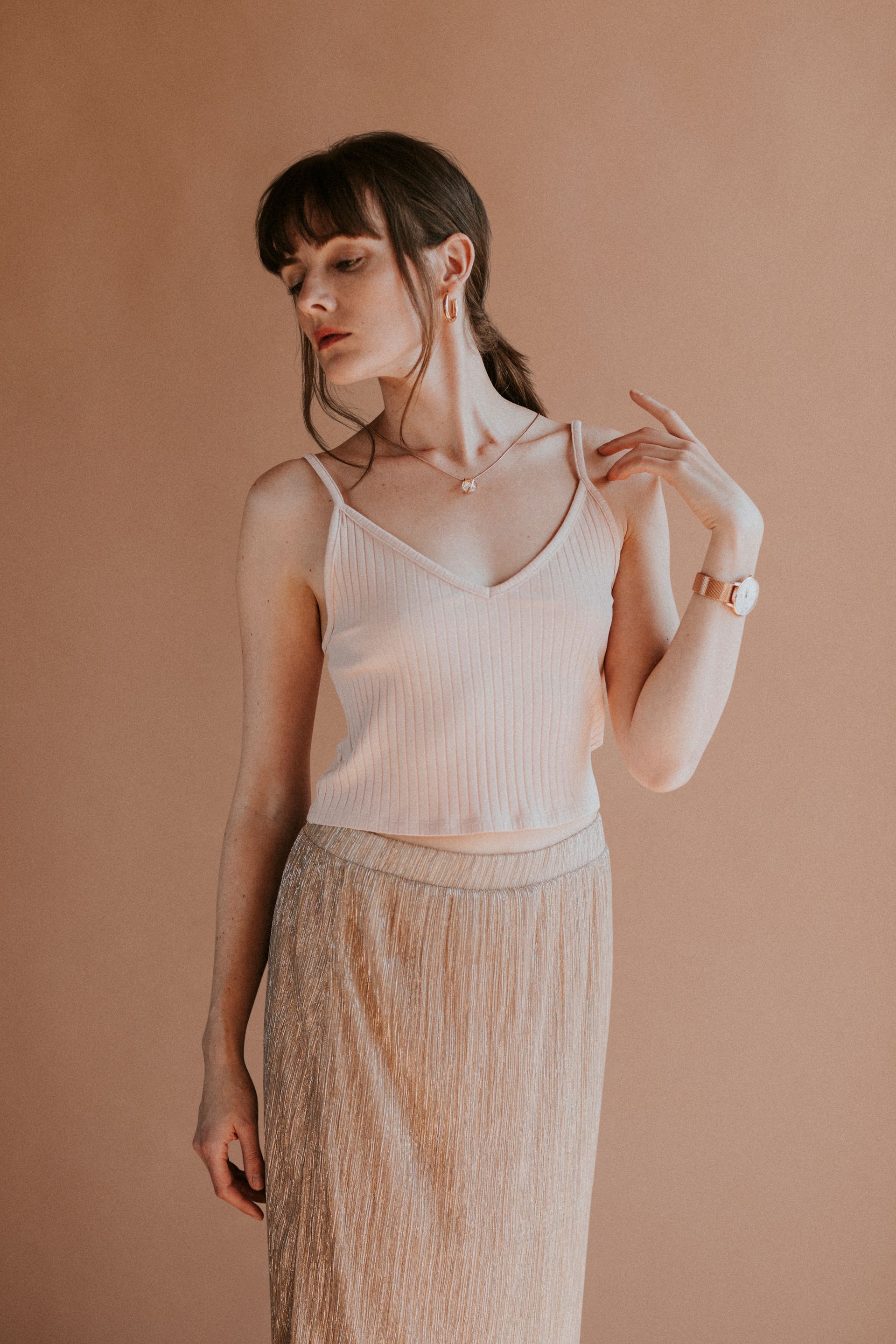 Woman in blush camisole and sequined skirt, gazing down, hand on shoulder, brown backdrop.