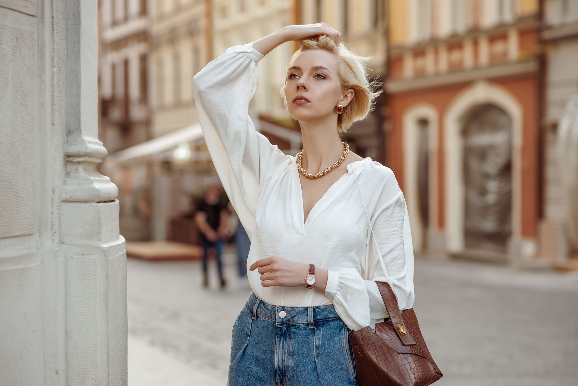 Woman in white top and jeans poses outdoors, city background, hand in hair.
