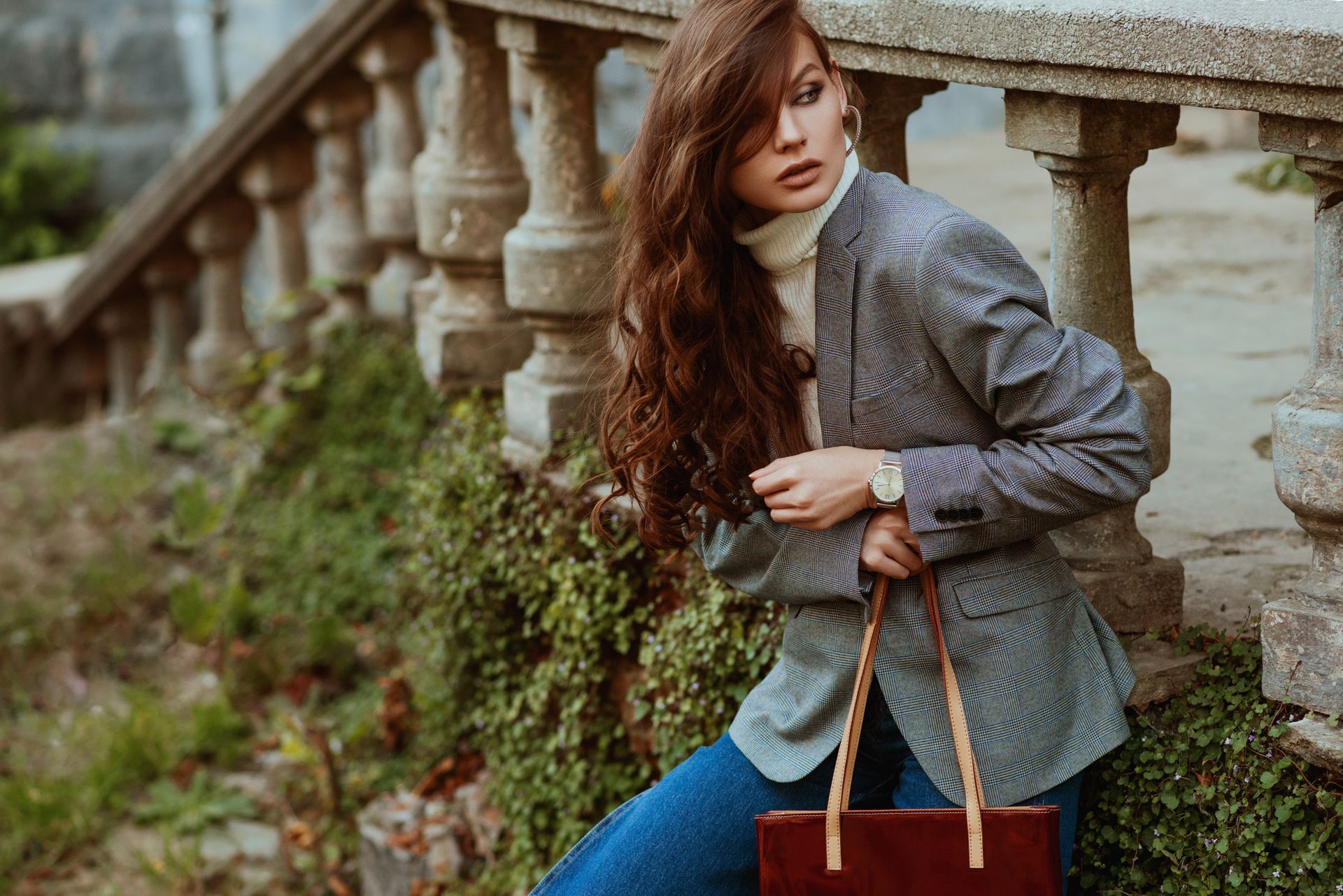 Woman in blazer and turtleneck, leaning against a stone railing, holding a brown bag.