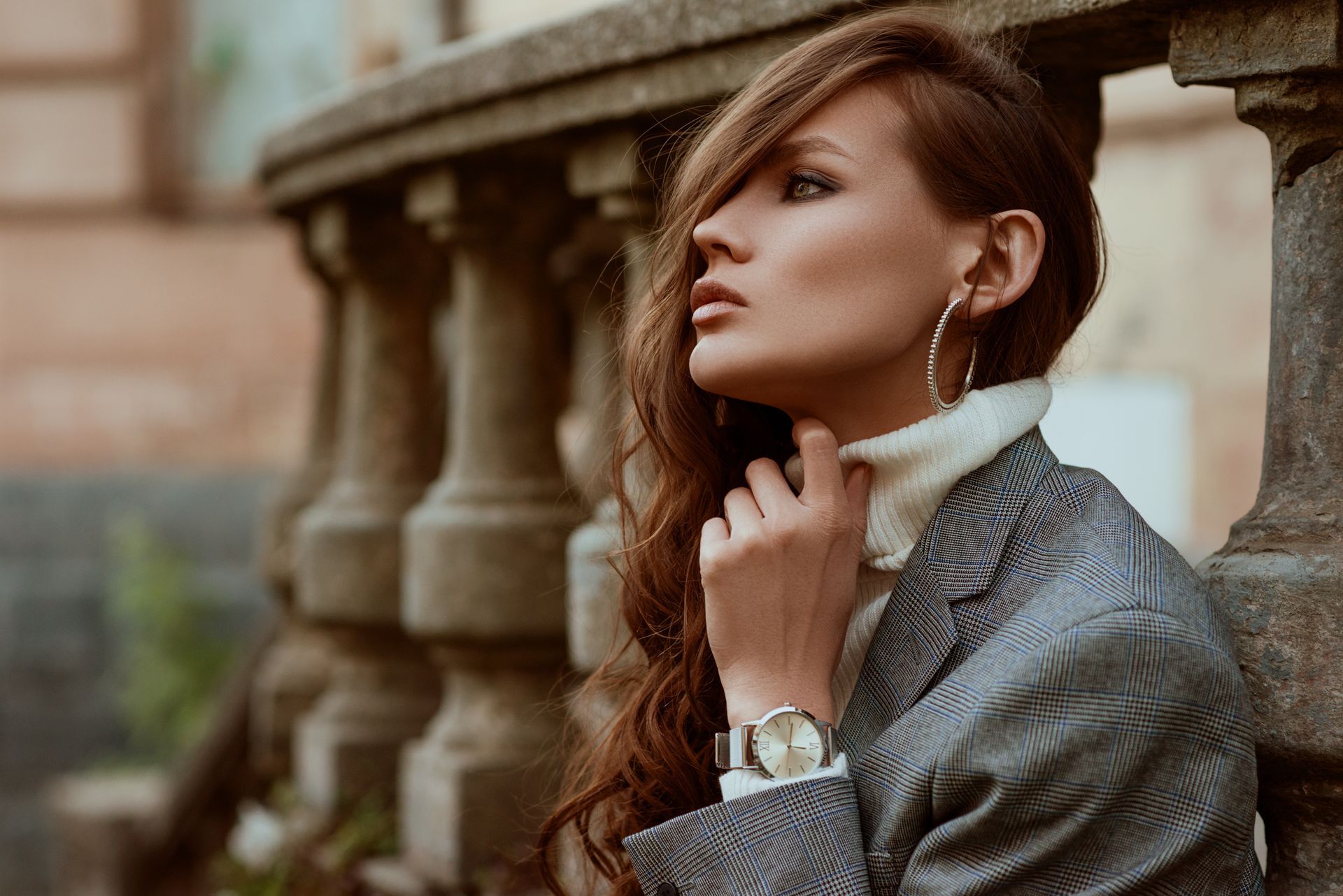 Woman in a blazer and turtleneck sweater, leaning against a stone railing, looking away, holding her neck.