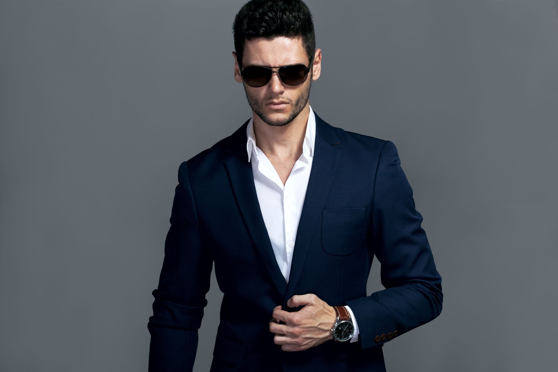 Man in navy suit, white shirt, sunglasses, buttoning jacket. Gray background.