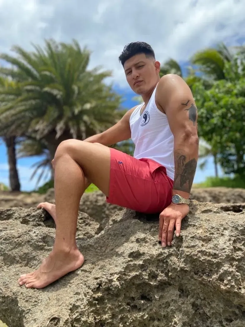 Man in white tank and red shorts sitting on a rock near palm trees.