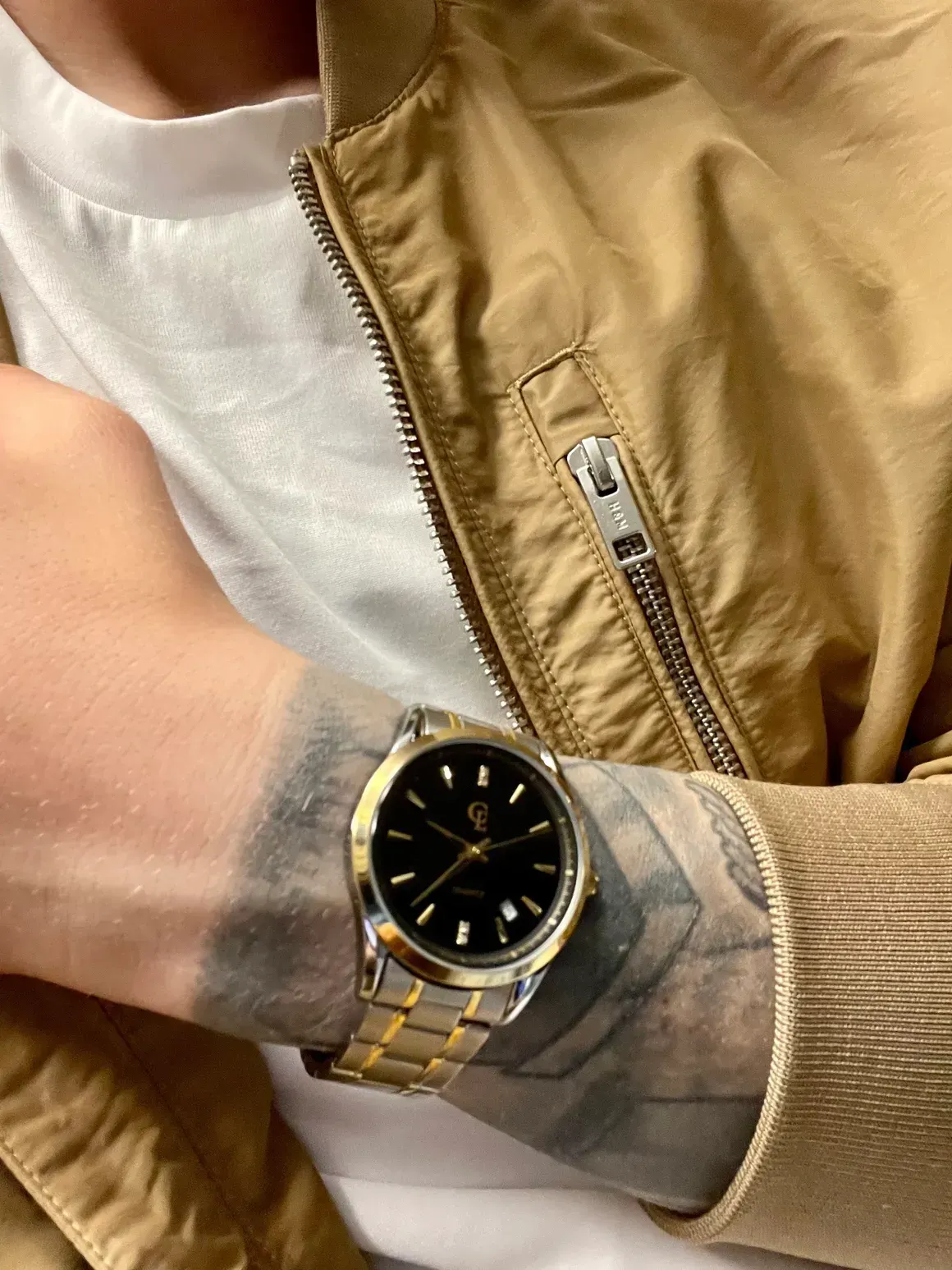 Wrist with gold and silver watch, black face, over tattooed arm, beige jacket.