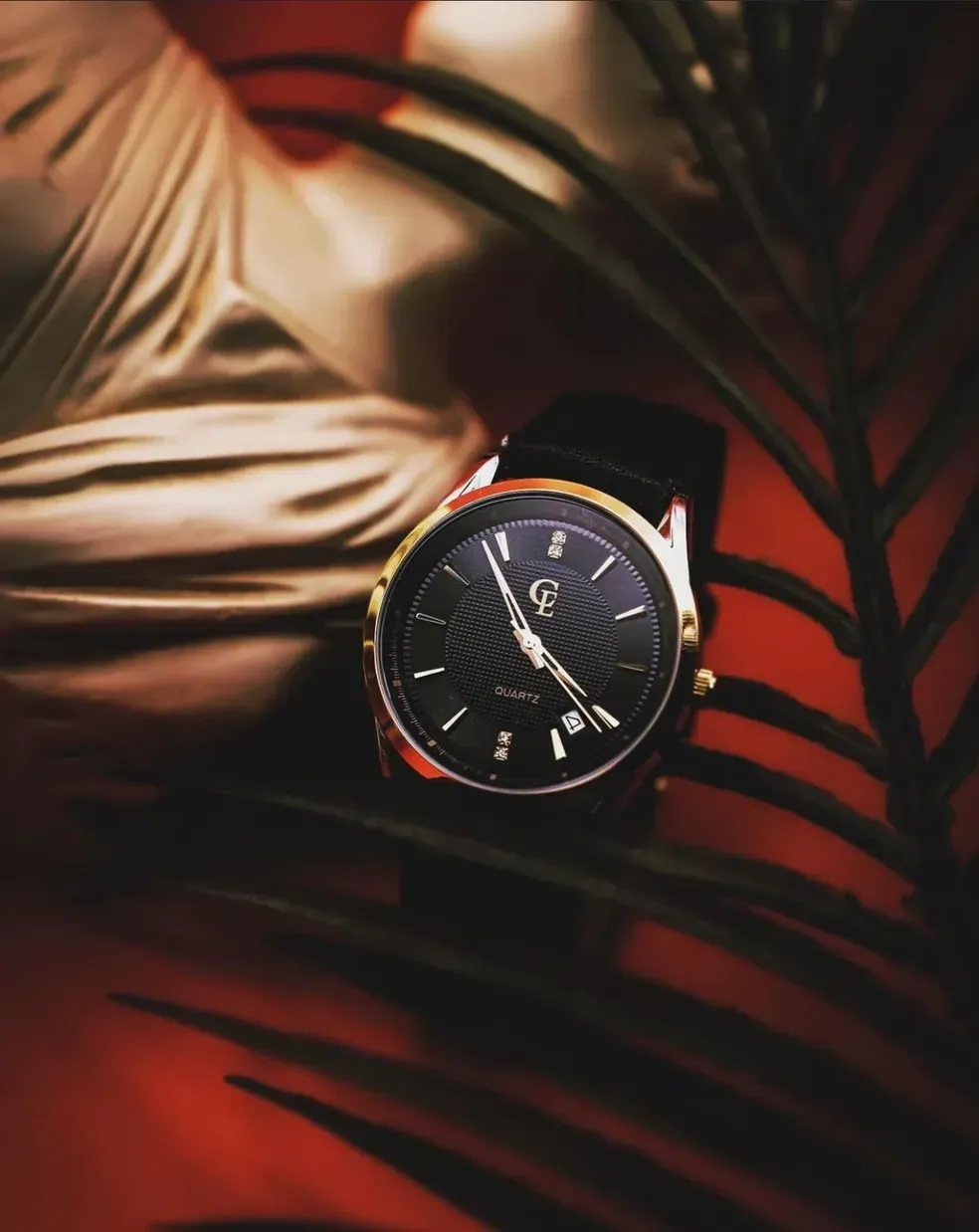 Black wristwatch with gold trim on a red surface, framed by palm leaves.
