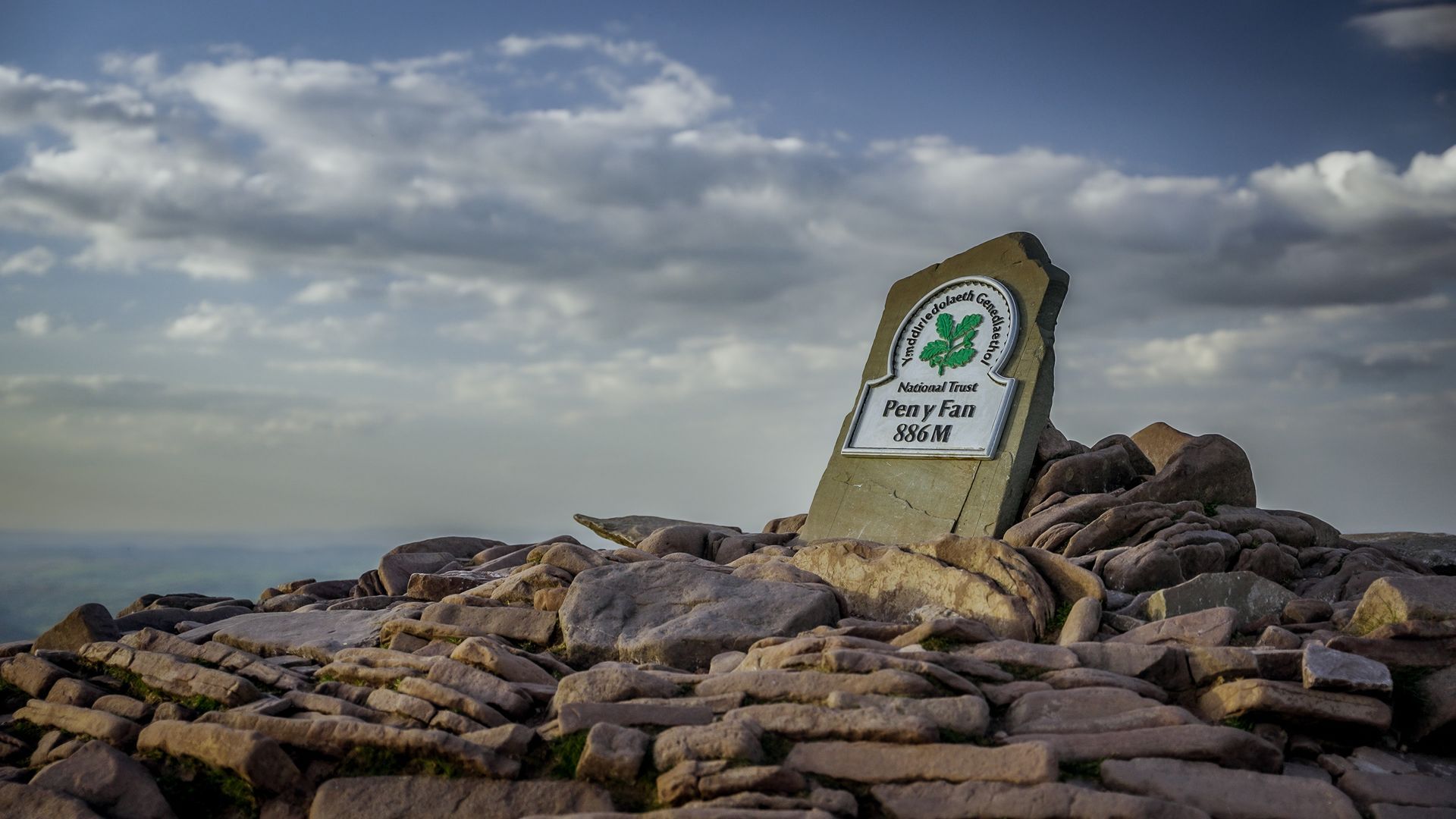 Stone marker on mountain peak, green emblem, rocky landscape, blue sky with clouds.