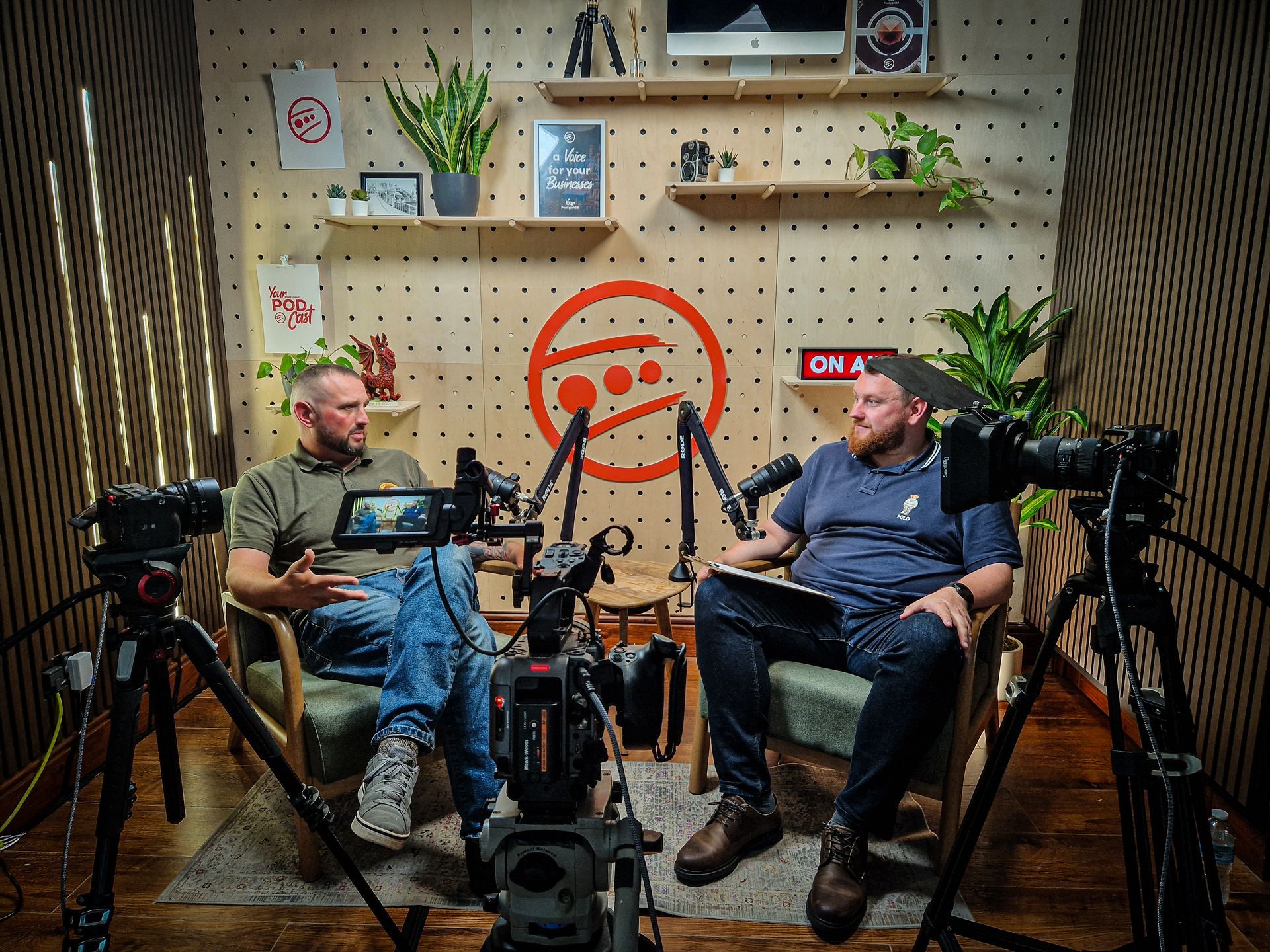 Two men seated in a podcast studio with microphones, laptops, plants, and a pegboard backdrop.