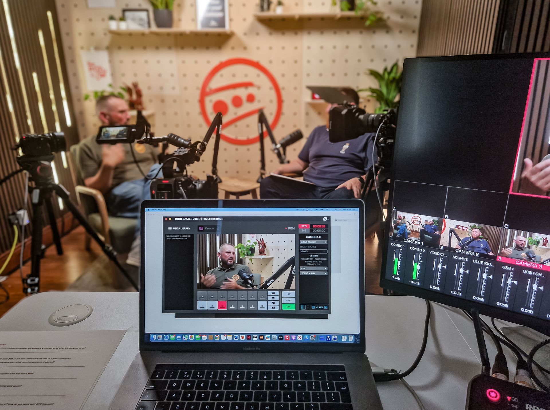 Podcast studio with two people recording, seen through a laptop and monitor setup
