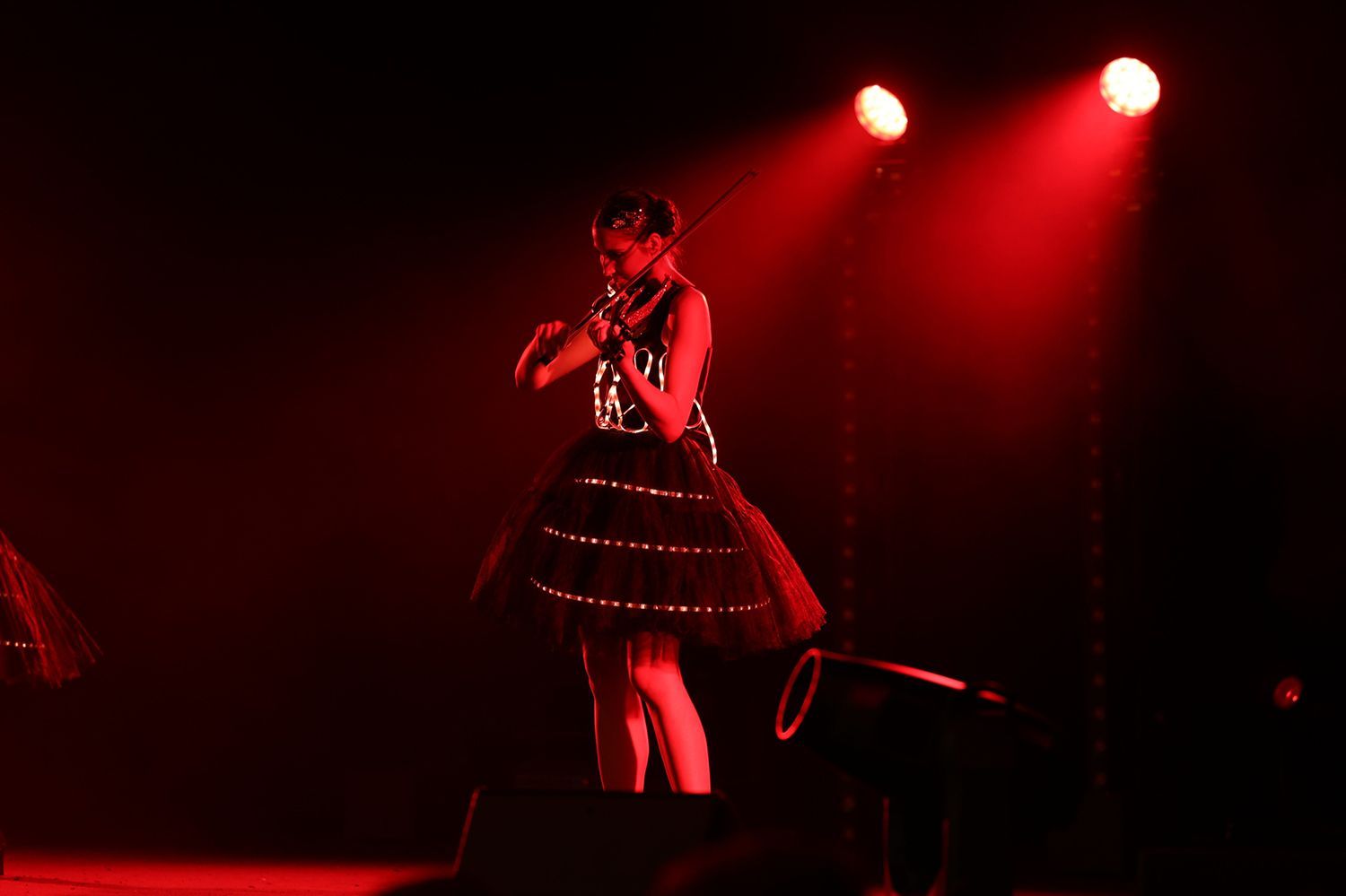 A performer on stage, lit by red spotlights, singing into a microphone. She wears a black dress with lights.