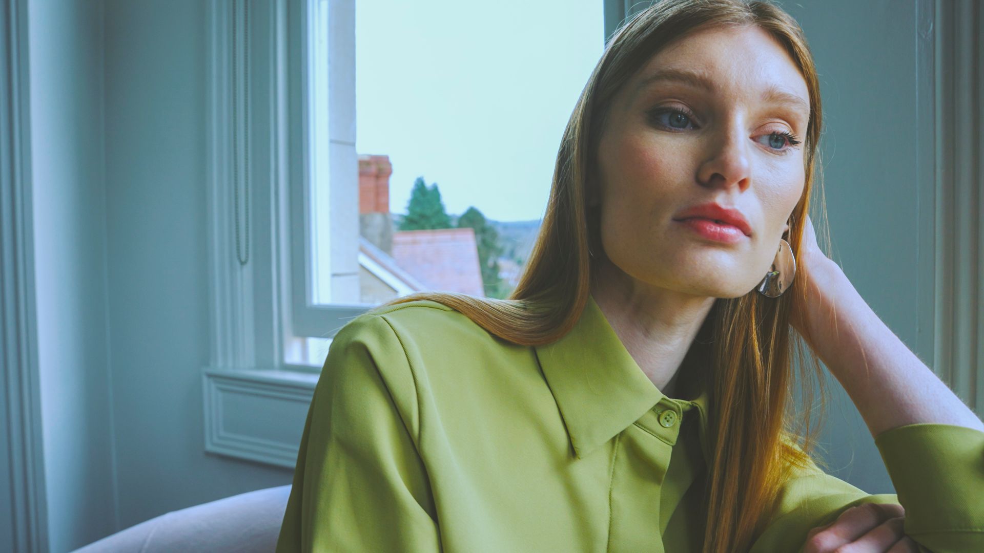 Woman in a green shirt, looking away from the camera, near a window with a view.