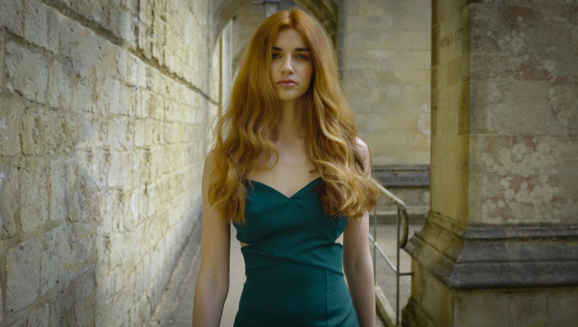 Woman with long red hair wearing a green dress walking through a stone archway.