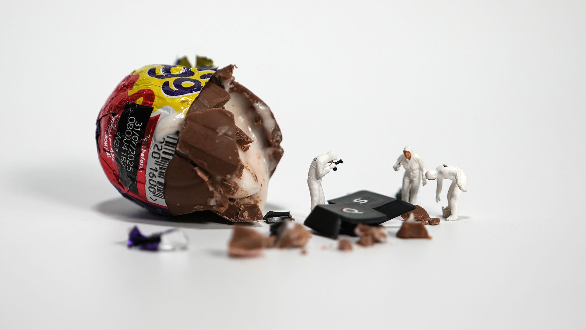 Miniature forensic investigators examining a partially eaten chocolate egg on a white surface.