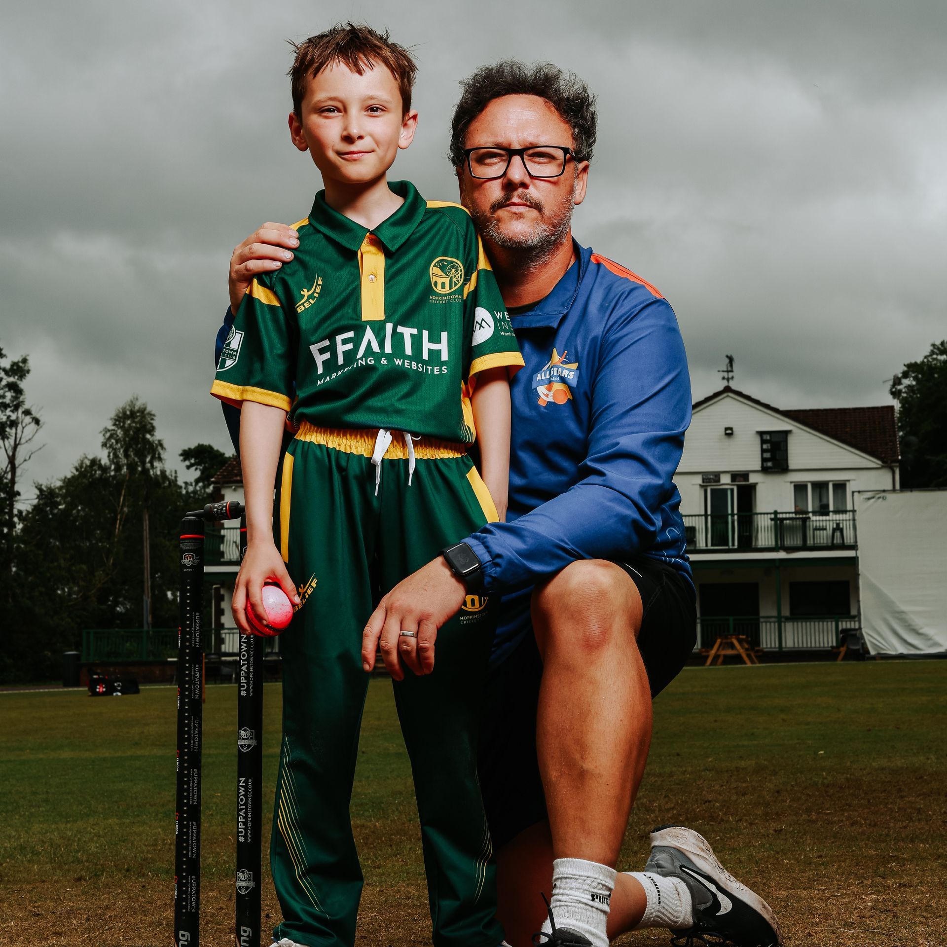 A boy in cricket attire with a coach, both on a cricket pitch. The coach has an arm around the boy's shoulder.