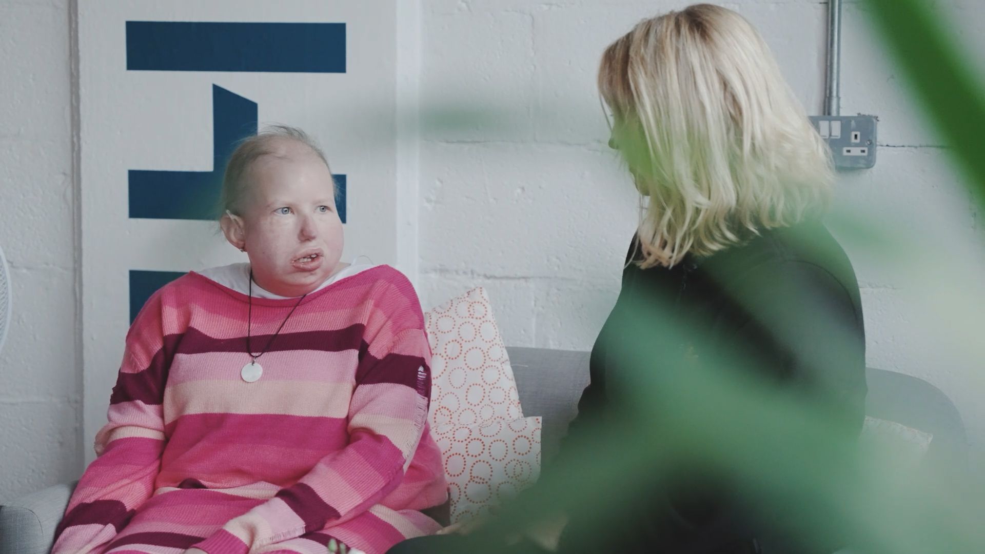 Woman in pink sweater talking with another blonde woman in a room with a partial white wall and plants.
