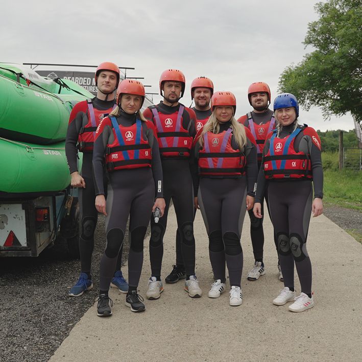 Group of people in wetsuits and life vests pose by rafts; outdoors.