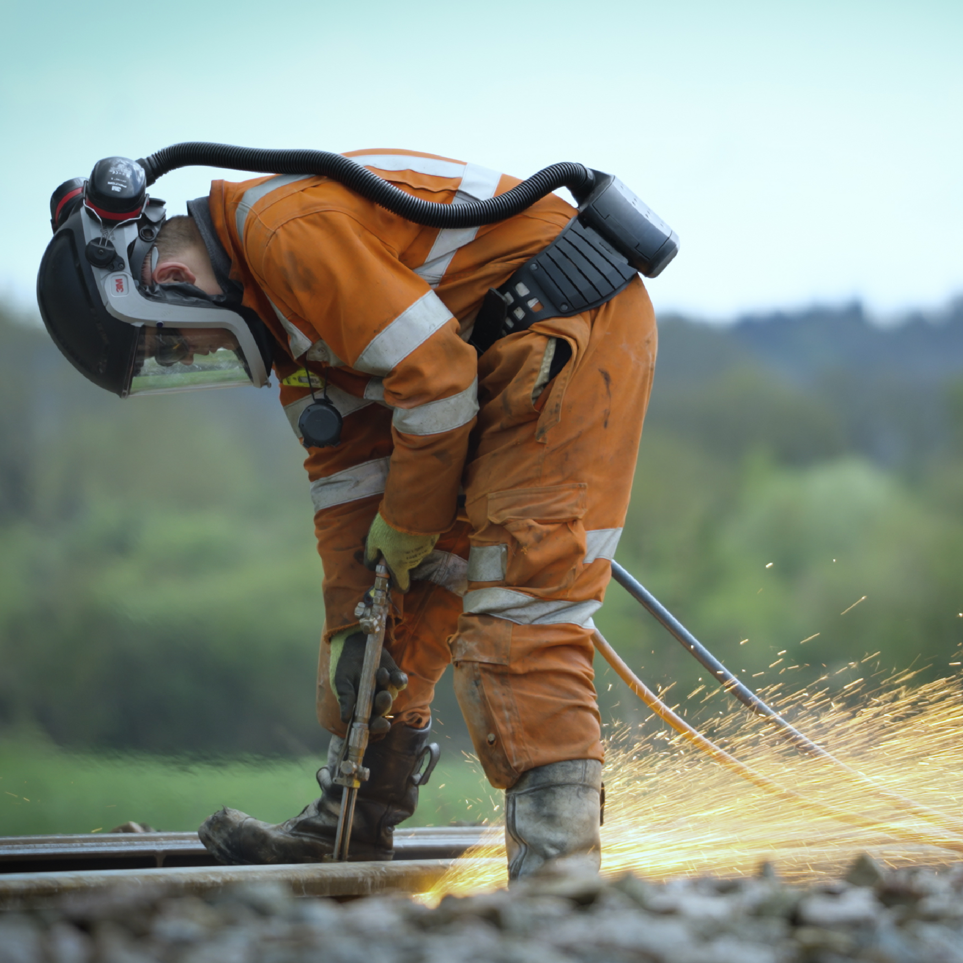 Rail worker in orange protective gear, grinding metal with sparks flying. Outdoors, green background.