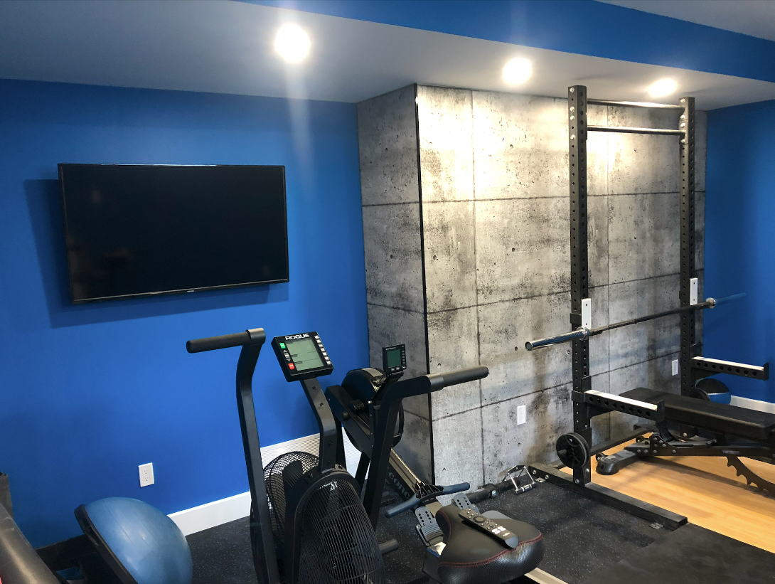 Home gym with blue walls, concrete accent wall, equipment like a rower, TV, and weight rack.