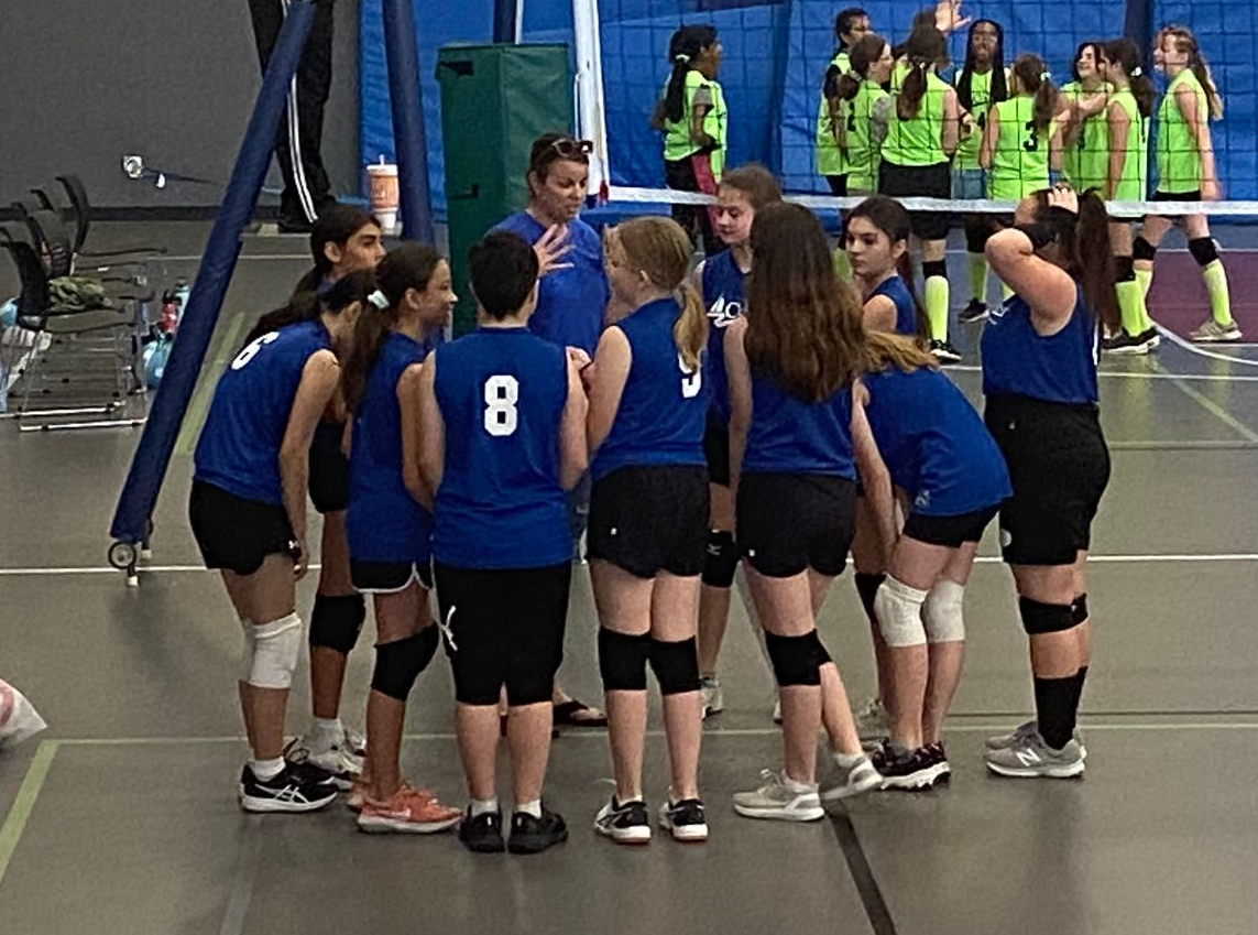 A youth volleyball team huddled around their coach who is giving them direction.