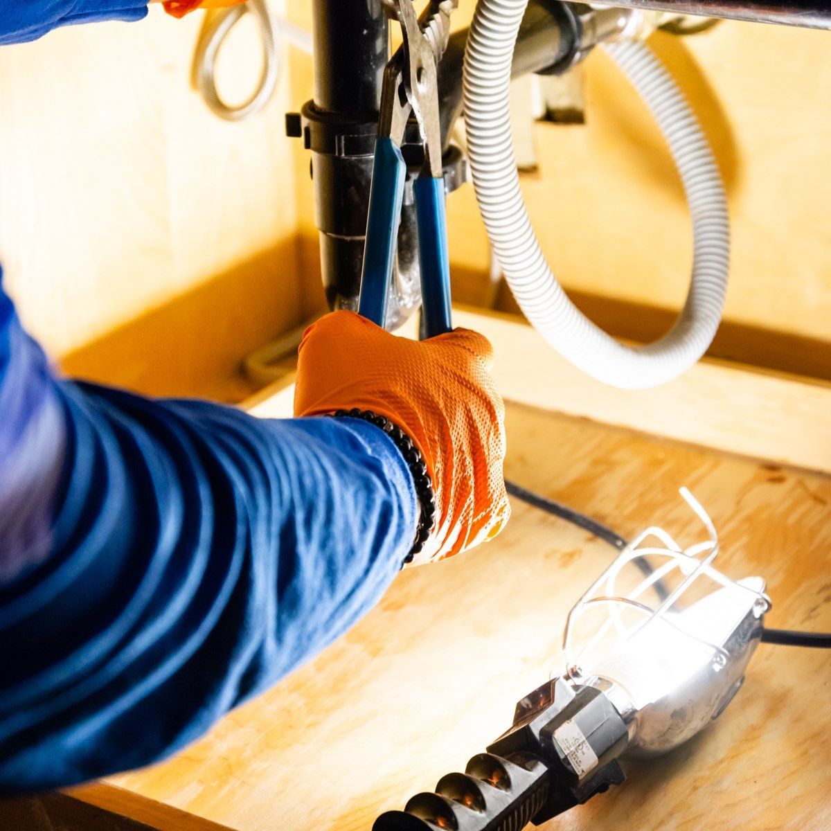 Plumber with orange gloves uses pliers under a sink, illuminated by a work light.