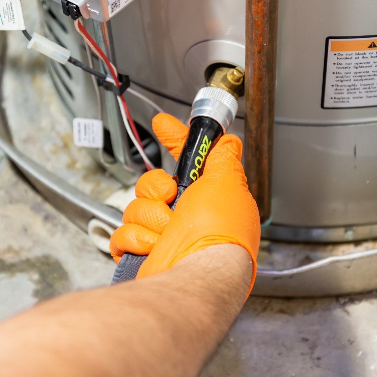 Person in orange gloves connecting a black hose to a water heater pipe in a basement.