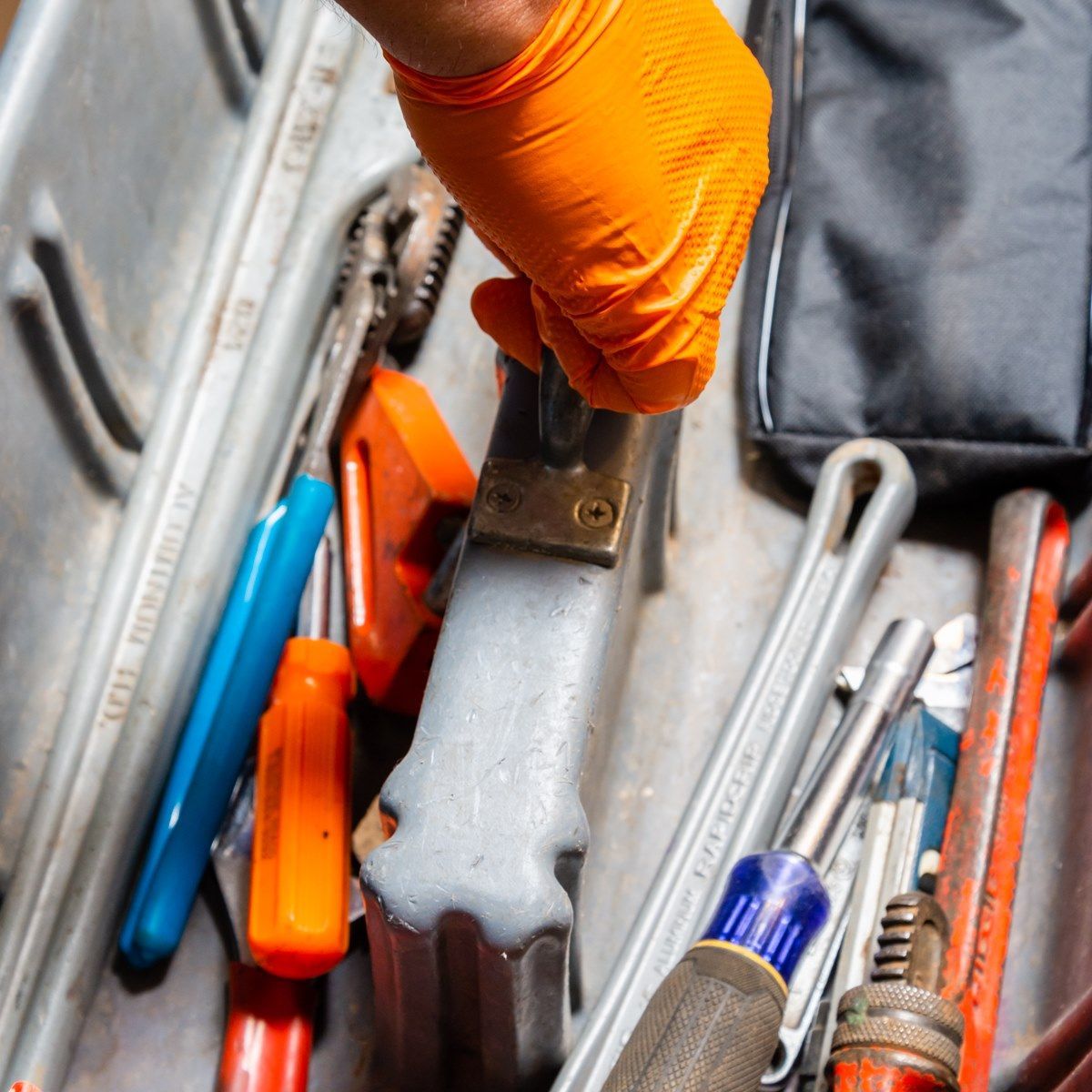 Hand in orange glove reaching for a tool in a toolbox filled with tools.