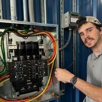 Technician beside open electrical panel, holding red and yellow wires in a metal utility room