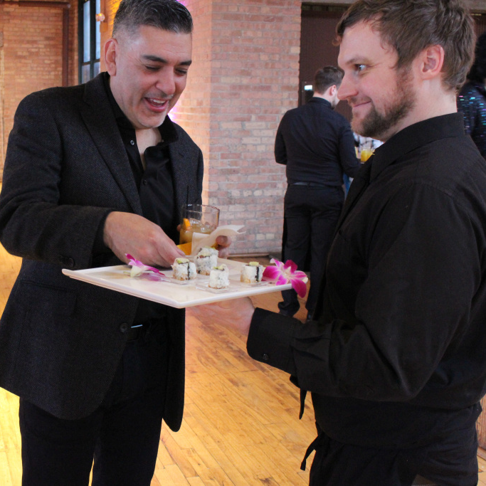 One man grabs a piece of sushi from a tray the other man is holding.