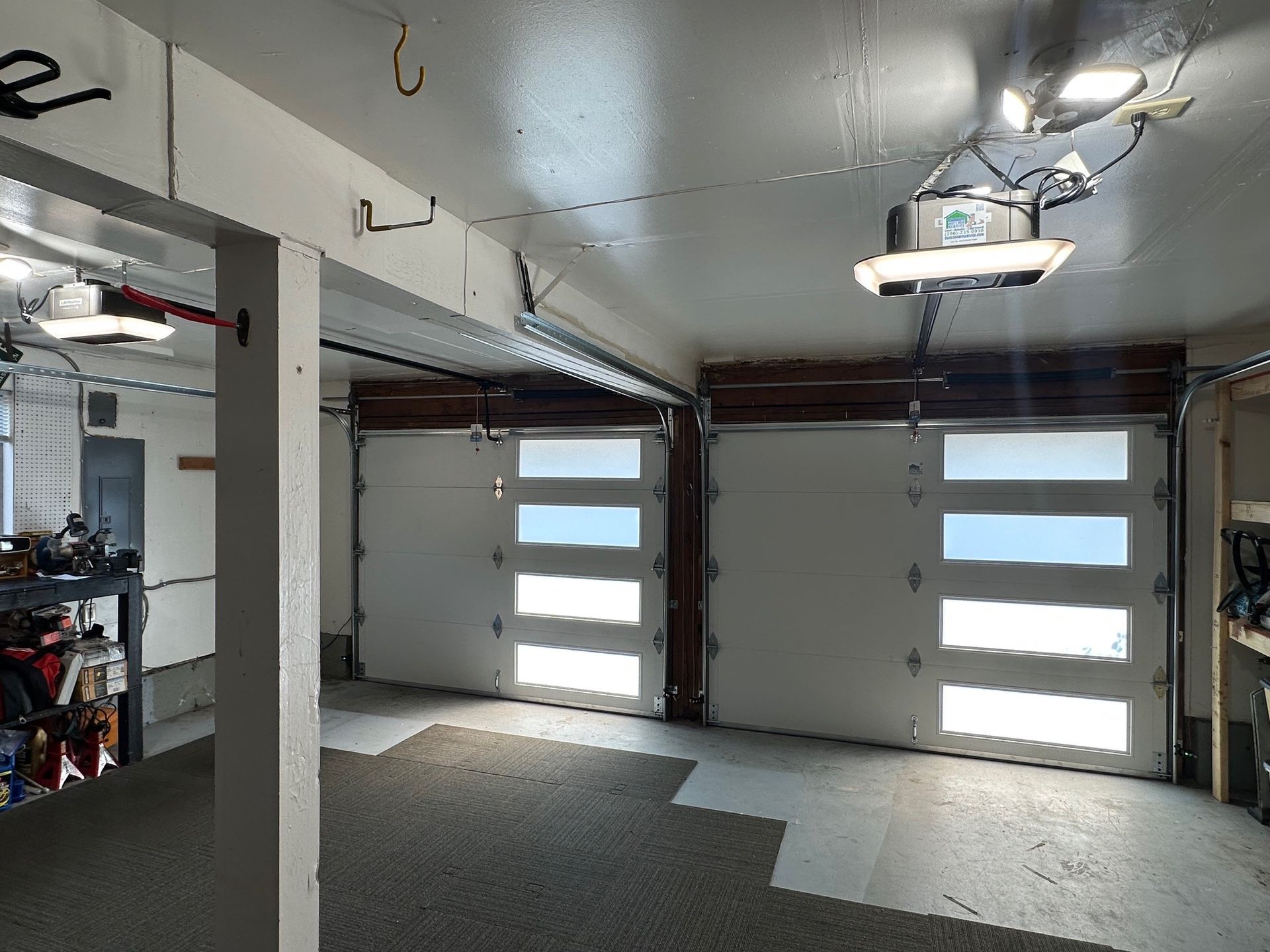 Garage interior with two white garage doors and a grey floor mat. Overhead lights illuminate the space.