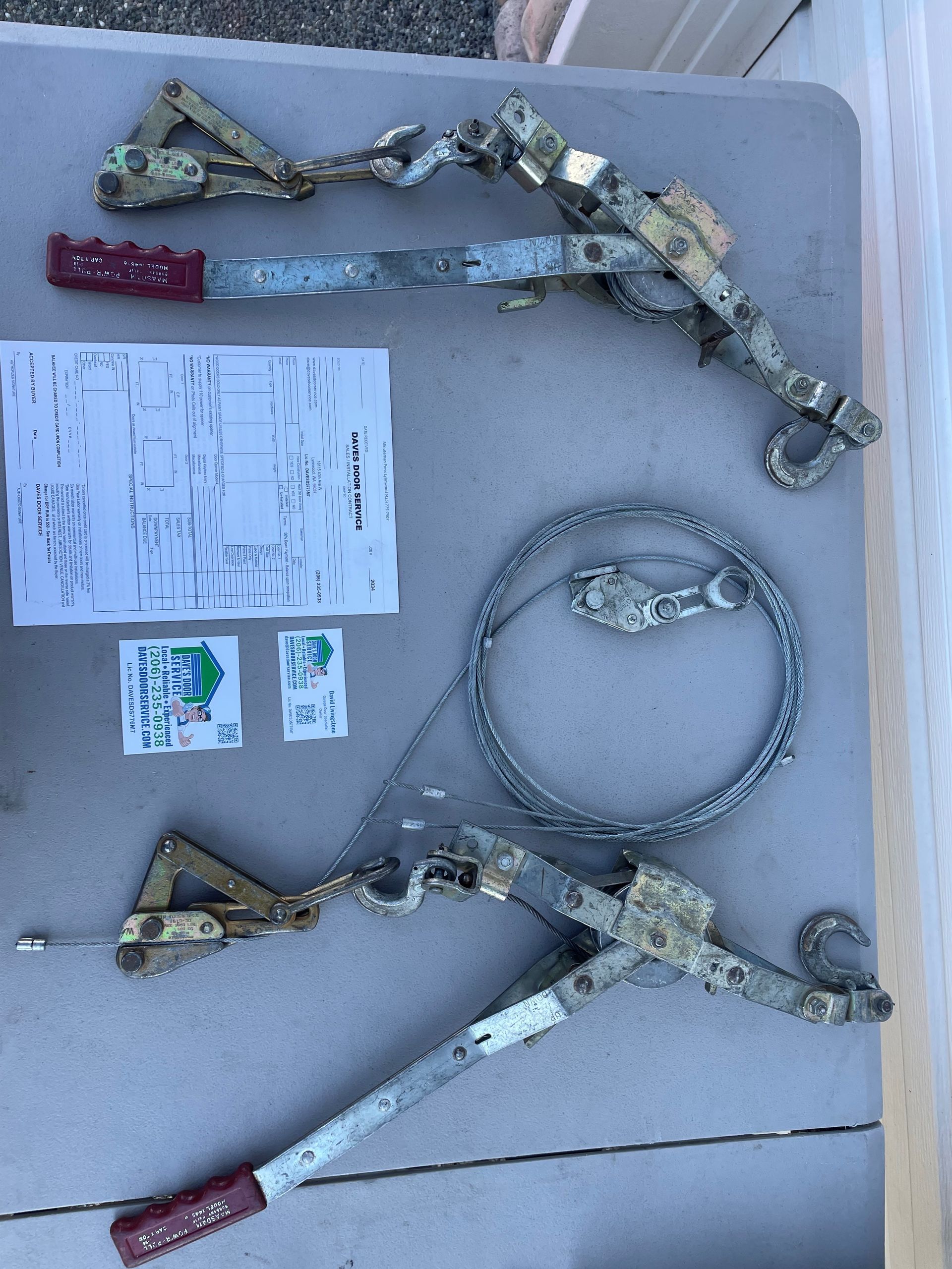 Two lever-type load binders, wire rope, and paper on a table. The binders have red handles.