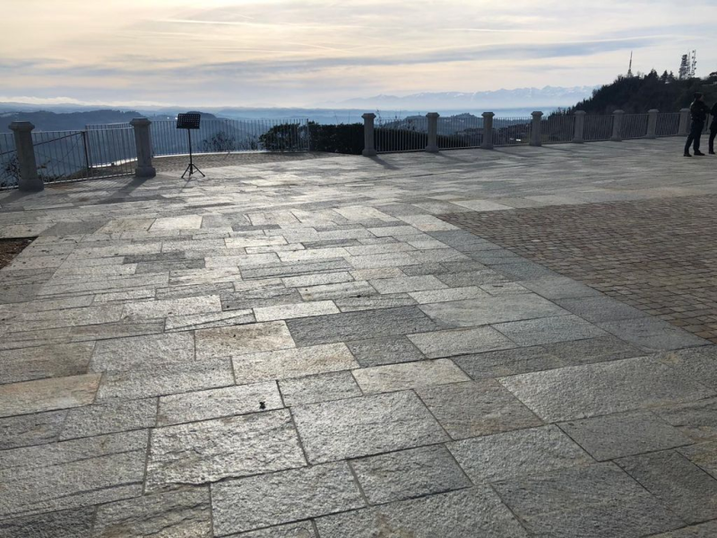 Terrazza in pietra con vista sulle colline in lontananza, sotto un cielo nuvoloso, con ringhiere e poche persone sullo sfondo.